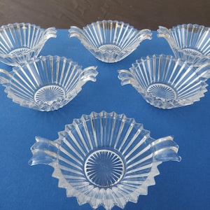 May include: Six clear glass serving dishes with a fluted design and scalloped edges. Each dish has two small handles and a circular pattern in the center. The dishes are arranged on a blue surface.