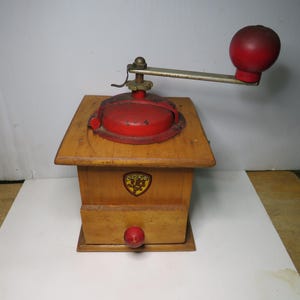 May include: A vintage wooden coffee grinder with a red handle and top. The grinder has a drawer and a metal crank. A gold and red emblem is on the front of the grinder.