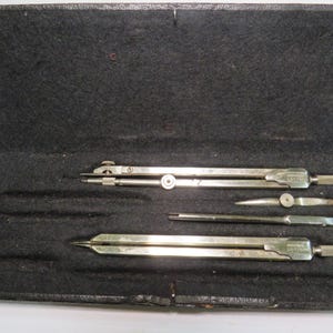 May include: An open black case containing a set of vintage drafting tools. The set includes a large compass, a smaller divider, and two other precision instruments. The tools are silver-colored and appear to be made of metal.