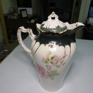 May include: Antique porcelain pitcher with a lid. The pitcher is white with a black rim and gold accents. It features a floral design in pink and green. The handle and lid are also detailed with gold trim.