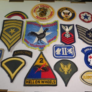 May include: A collection of vintage military patches, including those for the Marine Corps, Navy, and Army. Patches feature eagles, stars, and text like "Airborne" and "Hell on Wheels." Colors include blue, gold, red, and green.