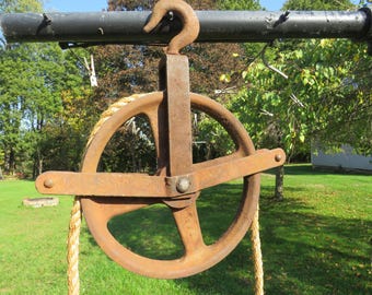 Puede incluir: Un antiguo sistema de polea de metal oxidado con una cuerda gruesa. La rueda de la polea es circular con un soporte en forma de cruz. El sistema está suspendido de una barra de metal negro con un gancho curvo. El fondo presenta hierba verde y árboles.