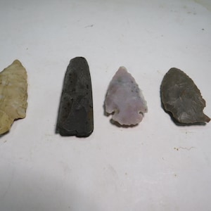 May include: Four ancient stone artifacts are displayed against a white background. The collection includes a light-colored arrowhead, a dark gray, elongated tool, a pale purple arrowhead, and a gray arrowhead.