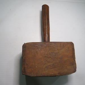 May include: A wooden mallet featuring a rectangular head with a visible grain pattern and a cylindrical handle attached to the center. The wood displays a medium brown tone with natural texture and signs of wear across the surface.
