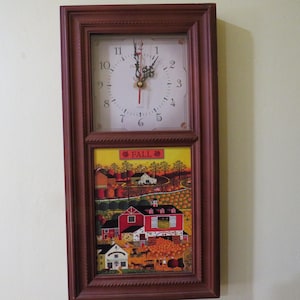 May include: A rectangular brown framed wall clock with a white clock face and a fall-themed landscape painting. The clock face has black numbers and hands. The painting depicts a red barn, houses, and trees.