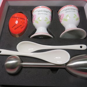 May include: A breakfast set in a box containing two white porcelain egg cups with pink floral patterns and the text Mevis Line, two white ceramic spoons, a stainless steel egg topper tool, and an orange plastic egg timer with black text.