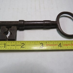 May include: An antique, dark metal key with a large, oval handle and a detailed bit. The key is approximately 5 inches long, lying next to a yellow measuring tape. The key's design suggests it is vintage.
