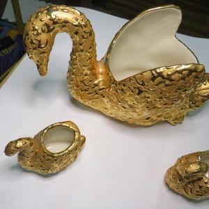 Peut inclure: Un centre de table décoratif en forme de cygne de couleur or avec une surface texturée. Le cygne a un intérieur blanc et est accompagné de deux petites pièces en forme de cygne, également de couleur or.
