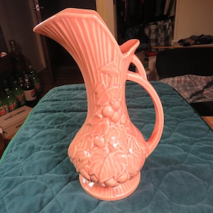 May include: A peach-colored ceramic pitcher with a tall, fluted neck and a curved spout. The body features a raised leaf and berry design. It has a large handle and sits on a round base. A decorative item.