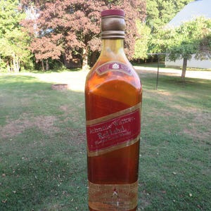 May include: A tall, amber-colored bottle of Johnnie Walker Red Label whiskey. The bottle has a red and gold label with the brand name and product information. The bottle is set against a backdrop of green grass and trees.