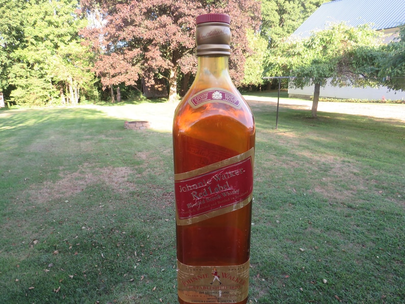 May include: A tall, amber-colored bottle of Johnnie Walker Red Label whiskey. The bottle has a red and gold label with the brand name and product information. The bottle is set against a backdrop of green grass and trees.