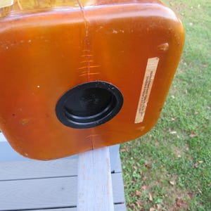 May include: An orange-brown plastic container with a black circular opening. The container has a rectangular shape with rounded corners. A label is affixed to the side. The container is resting on a light gray wooden surface.