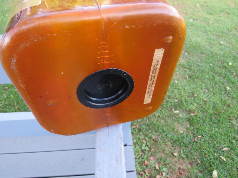 May include: An orange-brown plastic container with a black circular opening. The container has a rectangular shape with rounded corners. A label is affixed to the side. The container is resting on a light gray wooden surface.