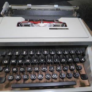 May include: A vintage Olivetti Lettera 31 typewriter. The typewriter has a light grey body with black keys and a black carriage. The keyboard layout includes numbers, letters, and symbols. The red ribbon is visible.