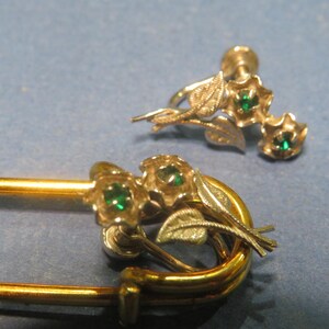 May include: A gold-tone safety pin brooch featuring a floral design with two small green stones and textured silver-tone leaves, accompanied by a matching screw-back earring with two green stones and silver-tone leaves.