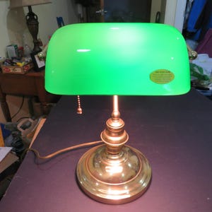 May include: A vintage-style desk lamp with a bright green glass shade. The lamp has a brass base and stem, and a pull chain for turning it on and off. The shade is curved and casts a soft, green glow. The lamp is designed for desk or table use.