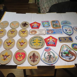 May include: A collection of Boy Scout patches, featuring various designs and colors. Patches include merit badges, service awards, and commemorative emblems. Some patches display text such as "Assistant Senior Patrol Leader" and "Uniform Award".