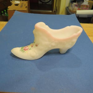 Peut inclure: Une chaussure en verre blanche de style vintage, avec un bord rose et des motifs floraux. La chaussure a un talon haut et présente un motif décoratif de roses roses et de feuilles vertes. Il s'agit probablement d'un objet de collection ou d'un élément décoratif.