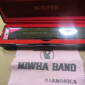 May include: A black harmonica in a red-lined case, with the text "HI-SUPER" on the case and harmonica. A pink cloth with "MIWHA BAND HARMONICA" is in front of the case.