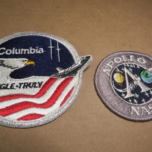 Peut inclure: Deux écussons brodés. L'un est rond avec un fond bleu, un aigle, une navette spatiale et les mots "Columbia" et "ENGLE-TRULY". L'autre est un cercle gris avec "APOLLO" et "NASA" et un "A" stylisé.