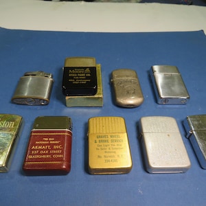 May include: A collection of vintage lighters in various shapes and finishes. The lighters include brands like Winston, Matchless, and Graves Wheel & Brake Service. Some lighters have text and logos, while others are plain metal.
