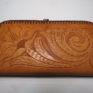 May include: A tan leather wallet with a floral design. The wallet features a detailed embossed flower and scroll pattern. The edges are stitched with dark brown thread, and it has a metal clasp closure.