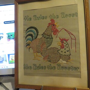 May include: Framed embroidered artwork featuring a rooster and hen with the text "He Rules the Roost" and "She Rules the Rooster". The rooster has a red comb and a green and blue tail. The hen has a red comb and is under a red barn.