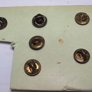May include: Five circular metal pin backs with a bronze-toned finish, each featuring a central loop mechanism for attachment. These pin backs are arranged on a light green cardstock backing, showing the reverse side of the pin hardware.