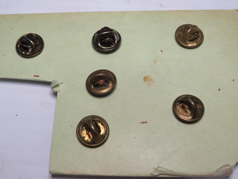 May include: Five circular metal pin backs with a bronze-toned finish, each featuring a central loop mechanism for attachment. These pin backs are arranged on a light green cardstock backing, showing the reverse side of the pin hardware.