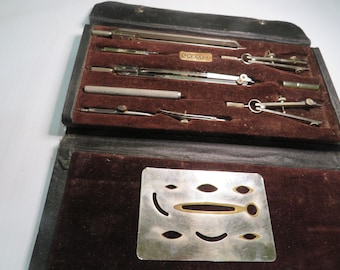 May include: An open, vintage drafting set in a black leather case with a dark brown velvet interior. The set includes several silver-colored compasses, dividers, and a stencil. A small, rectangular brass plate with text is visible.
