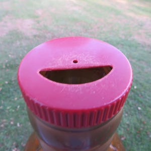 May include: Close-up of a red plastic bottle cap with a slot and a small hole. The cap is on a brown bottle, with a green grassy background.