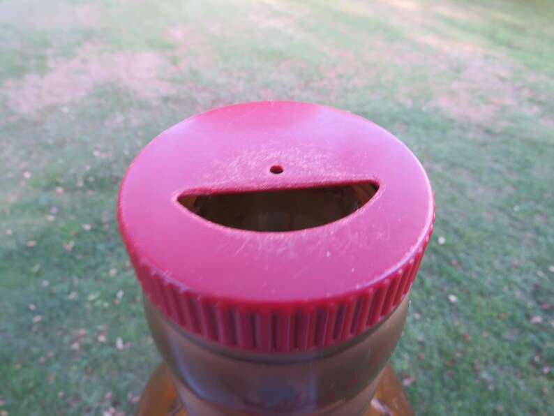 May include: Close-up of a red plastic bottle cap with a slot and a small hole. The cap is on a brown bottle, with a green grassy background.