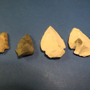 May include: Four ancient stone arrowheads of varying sizes and colors are arranged on a blue surface. The arrowheads range in color from tan to gray and off-white, showcasing their unique textures and shapes.