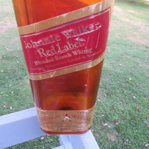 May include: A large, amber-colored bottle with a red and gold label. The label reads "Johnnie Walker Red Label Blended Scotch Whisky." The bottle is displayed outdoors, with a blurred background of grass and a white railing.