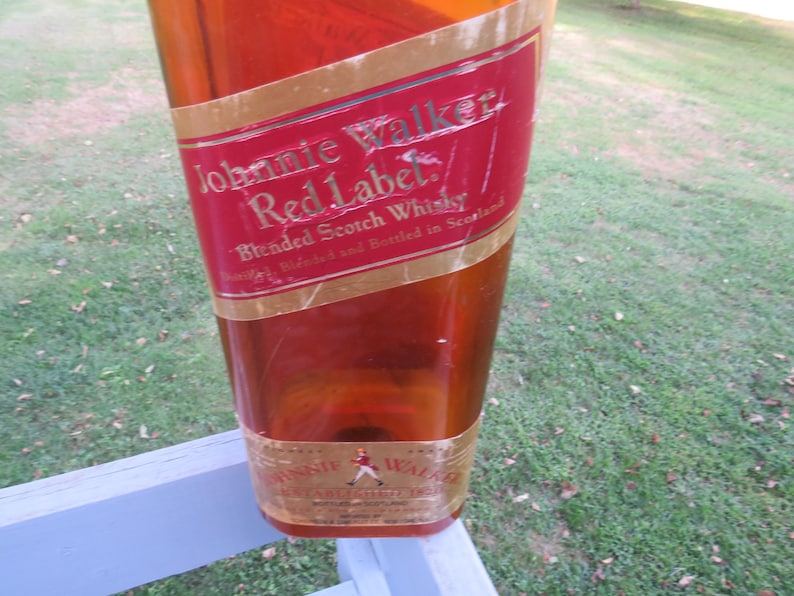 May include: A large, amber-colored bottle with a red and gold label. The label reads "Johnnie Walker Red Label Blended Scotch Whisky." The bottle is displayed outdoors, with a blurred background of grass and a white railing.
