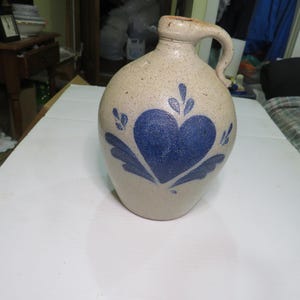 May include: A tan stoneware jug with a blue heart and leaf design. The jug has a handle and a spout. The heart is in the center of the jug, with leaf-like designs around it. The jug is likely used for storing liquids.