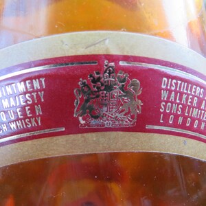 May include: Close-up of a vintage Scotch whisky bottle label. The label is red with gold lettering and a gold emblem. Text includes "By Appointment to Her Majesty The Queen Scotch Whisky" and "Distillers John Walker and Sons Limited London."
