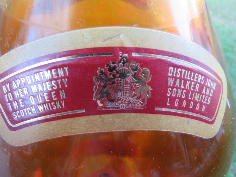 May include: Close-up of a vintage Scotch whisky bottle label. The label is red with gold lettering and a gold emblem. Text includes "By Appointment to Her Majesty The Queen Scotch Whisky" and "Distillers John Walker and Sons Limited London."