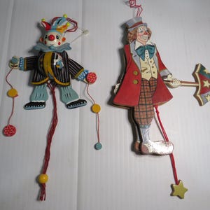 May include: Two wooden clown ornaments. One clown wears a striped jacket and a colorful hat. The other clown wears a red coat and holds a decorative umbrella. Both have strings attached for hanging.