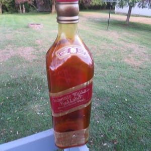 May include: A large, decorative bottle of Johnnie Walker Red Label Scotch Whisky. The bottle is mostly red and gold with a red label and gold lettering. The bottle is sitting on a blue surface with a green grassy background.