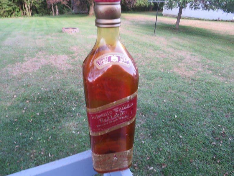 May include: A large, decorative bottle of Johnnie Walker Red Label Scotch Whisky. The bottle is mostly red and gold with a red label and gold lettering. The bottle is sitting on a blue surface with a green grassy background.