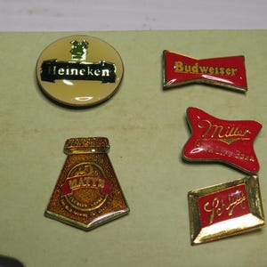 May include: A collection of six metal enamel pins featuring various beer brand logos including Heineken, Budweiser, Miller, and Matt's. The pins are shaped in circles, rectangles, and custom forms with red, gold, and black colors and metallic trim.