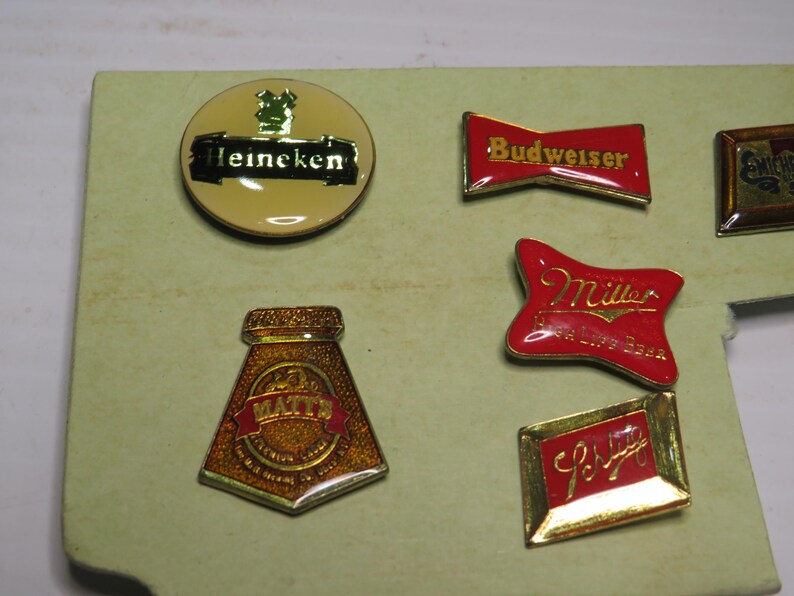 May include: A collection of six metal enamel pins featuring various beer brand logos including Heineken, Budweiser, Miller, and Matt's. The pins are shaped in circles, rectangles, and custom forms with red, gold, and black colors and metallic trim.