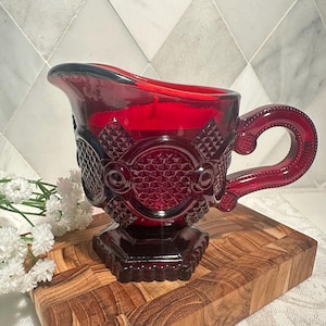 May include: A ruby red glass creamer with a decorative pattern and a handle. The creamer sits on a square base and has a spout for pouring. The creamer is displayed on a wooden cutting board.