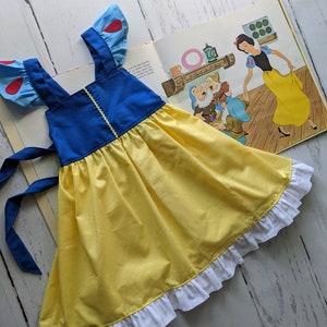 Puede incluir: Un vestido amarillo y azul con mangas en forma de coraz&oacute;n rojo y un ribete de volantes blancos. El vestido tiene un lazo azul para atar en la espalda. El vestido est&aacute; colocado sobre una superficie de madera blanca.