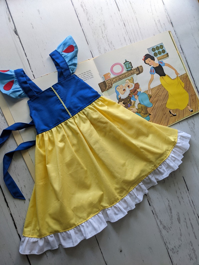 Puede incluir: Un vestido amarillo y azul con mangas en forma de coraz&oacute;n rojo y un ribete de volantes blancos. El vestido tiene un lazo azul para atar en la espalda. El vestido est&aacute; colocado sobre una superficie de madera blanca.