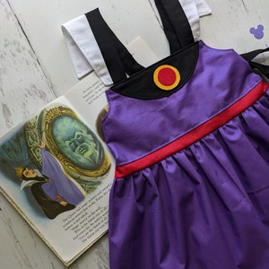 Op de afbeelding: Paarse jurk met zwarte bandjes, een rode tailleband en een gele en rode cirkel op het lijfje. De jurk ligt op een wit houten oppervlak met een boek en een paar rode Mickey Mouse oren.