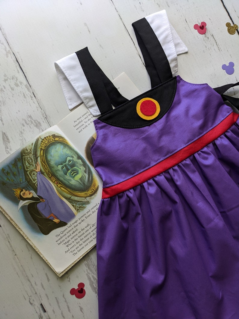 Op de afbeelding: Paarse jurk met zwarte bandjes, een rode tailleband en een gele en rode cirkel op het lijfje. De jurk ligt op een wit houten oppervlak met een boek en een paar rode Mickey Mouse oren.