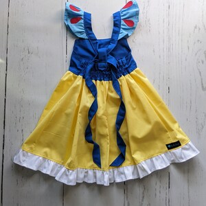 Puede incluir: Un vestido amarillo y azul con un ribete de volantes blancos. El vestido tiene un corpi&ntilde;o azul con detalles rojos y un lazo azul para atar en la espalda. El vestido est&aacute; hecho para un ni&ntilde;o.