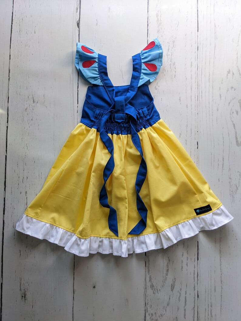 Puede incluir: Un vestido amarillo y azul con un ribete de volantes blancos. El vestido tiene un corpi&ntilde;o azul con detalles rojos y un lazo azul para atar en la espalda. El vestido est&aacute; hecho para un ni&ntilde;o.